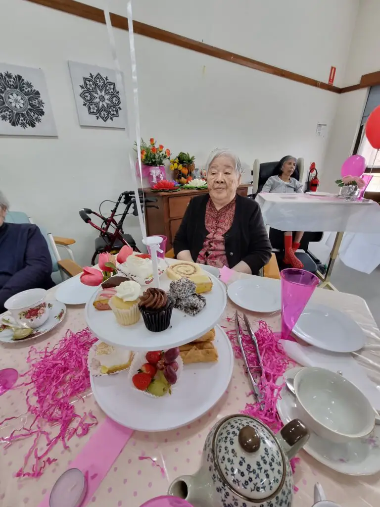 elderly woman sat in a table filled with small cakes, cupcakes and other sweet treats