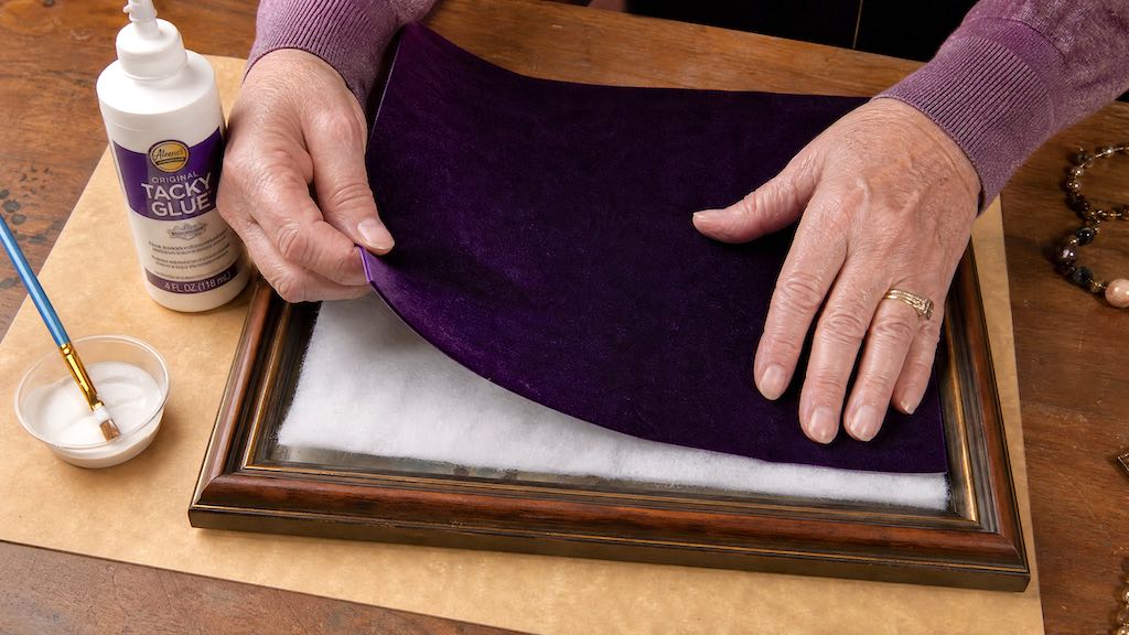 Dark velvet fabric being placed over cotton batting on a picture frame glass panel to create the jewelry art base