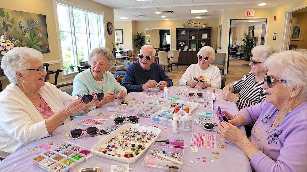 Seniors in an aged care setting decorating custom sunglasses with colorful gems, stickers, and craft supplies during a fun group activity.
