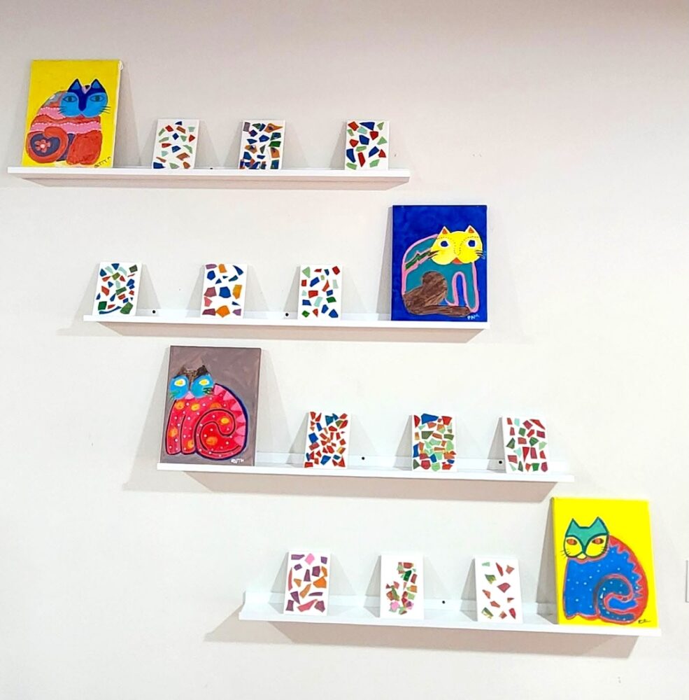 Finished lasagne mosaic canvases displayed on white wall shelves alongside colourful painted cat artworks