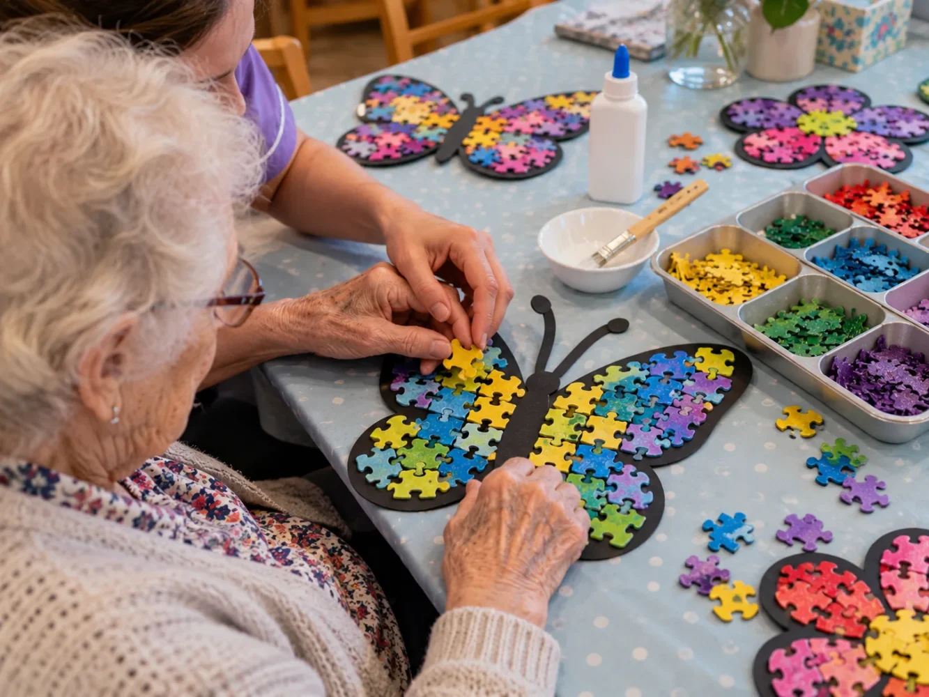 Carer giving hand-under-hand support to a resident placing puzzle pieces on a black card butterfly